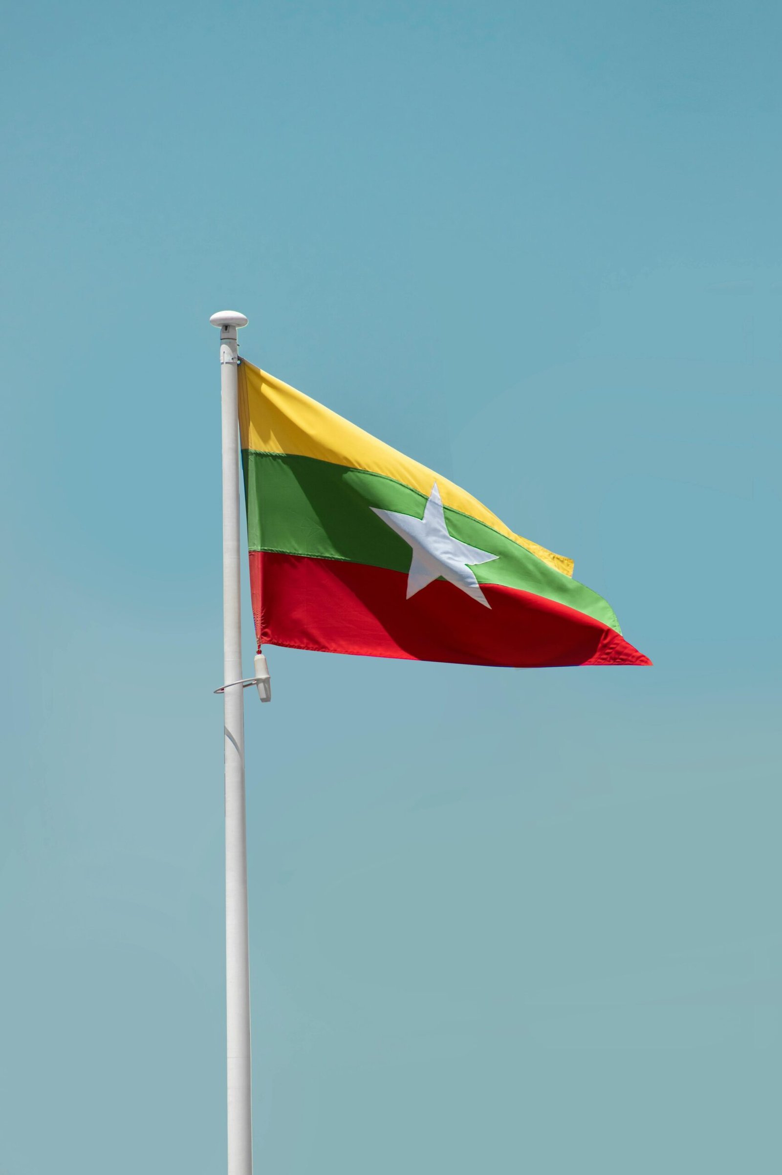The national flag of Myanmar waving on a flagpole against a clear blue sky.